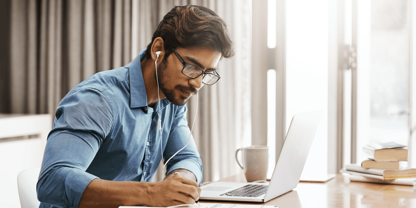 person sitting at a table at home in front of a laptop writing on a piece of paper wearing earbuds
