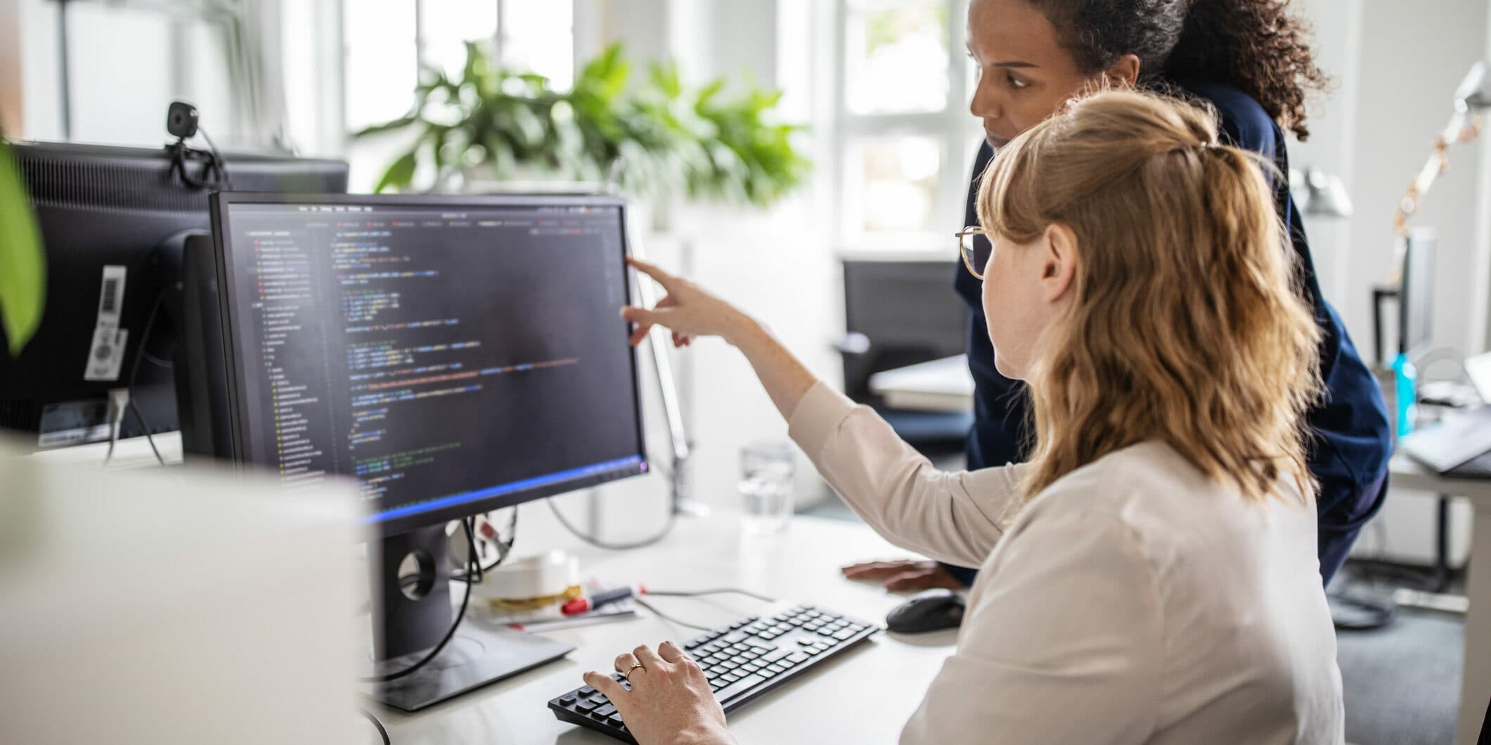 one person sitting at a desk pointing to a computer screen while another person looks at the screen