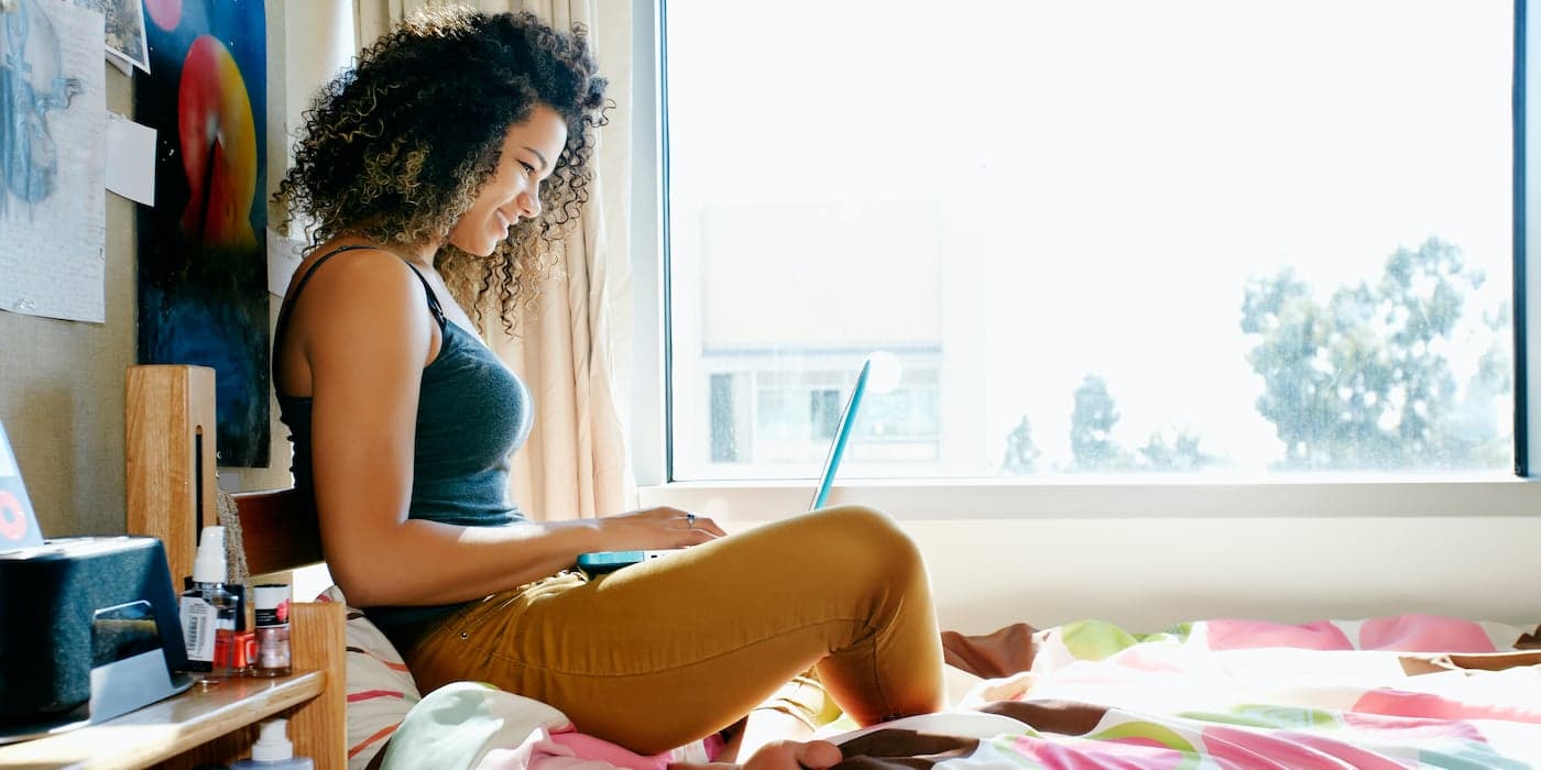 college student sitting on a dorm room bed working on a laptop