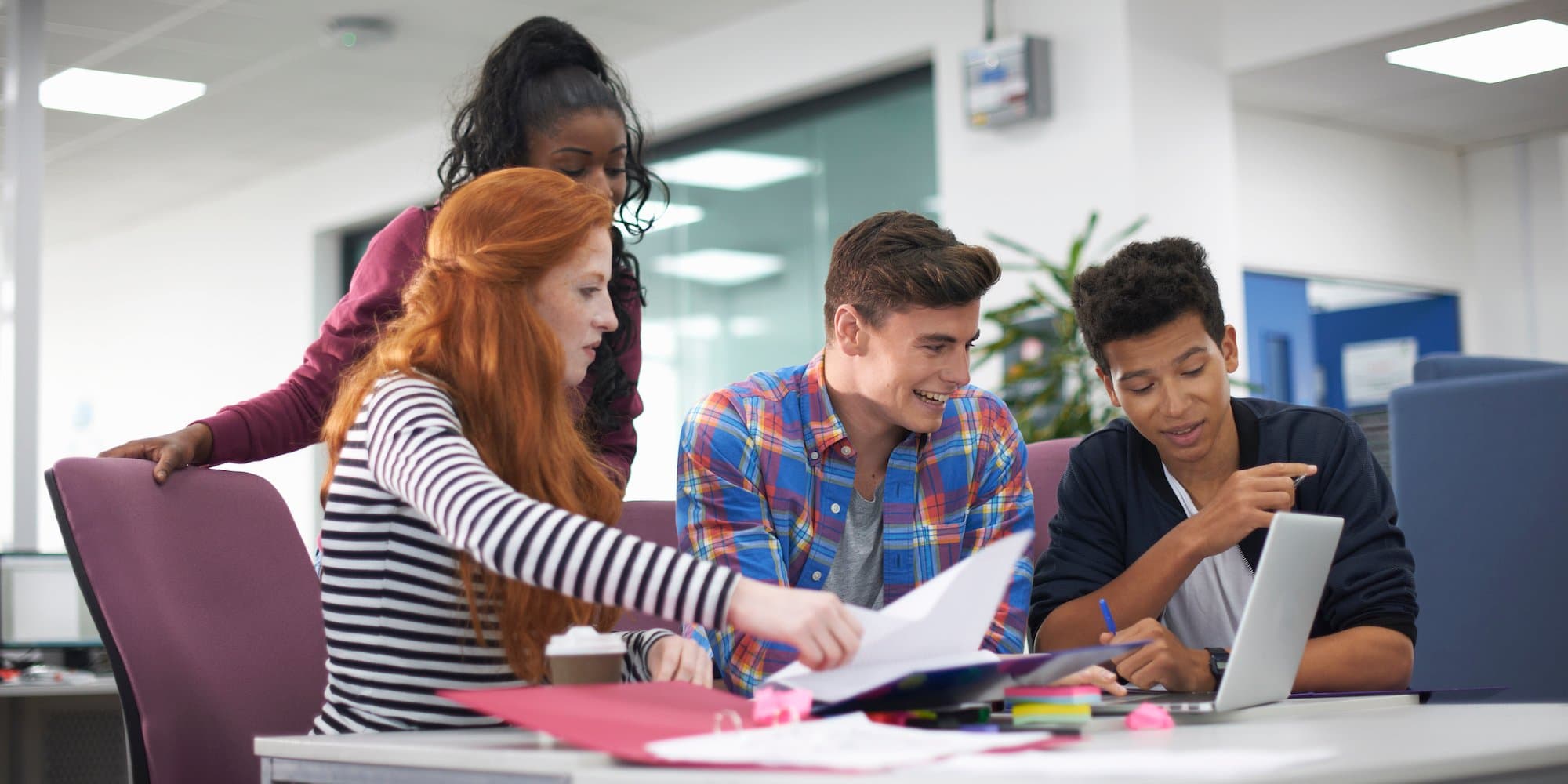group of college students crowded around table in library (three sitting, one standing) looking at laptop and papers