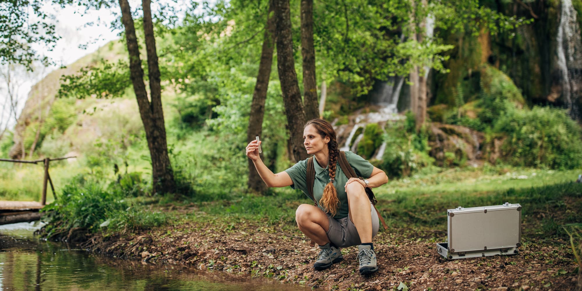 person crouching next to a stream in the forest, looking at a water sample in a test tube, with a case of supplies open next to them on the bank