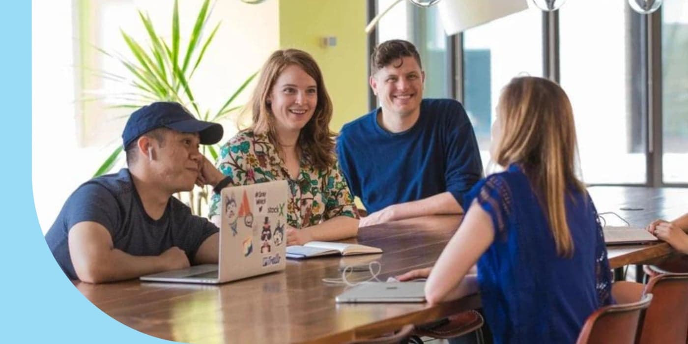 Four people gathered around a long table in an office space. The person on the far left wears a baseball cap and has an open laptop in front of them. The person on the far right sits on the opposite side of the table and has their back to the camera.