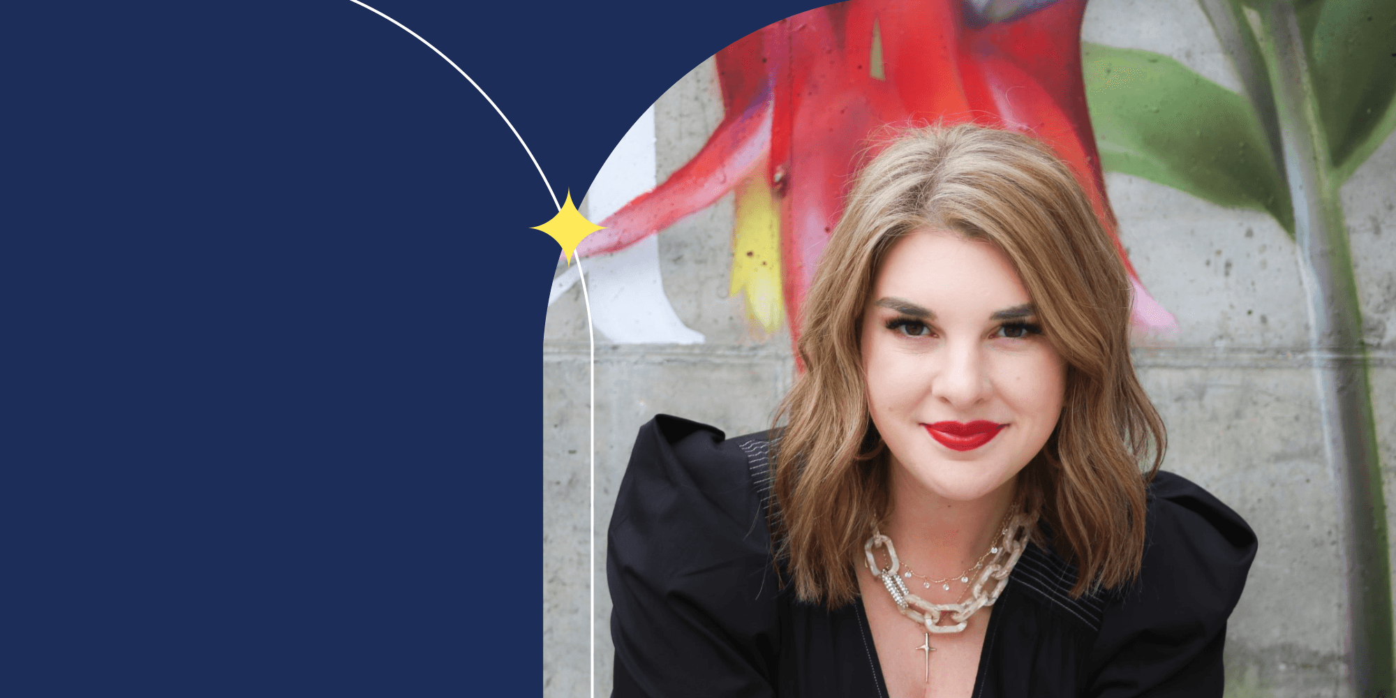 A person facing the camera; they are wearing red lipstick, a chunky necklace, and a black blouse. They have shoulder-length hair. Behind them, a wall painted with a flower can be seen.