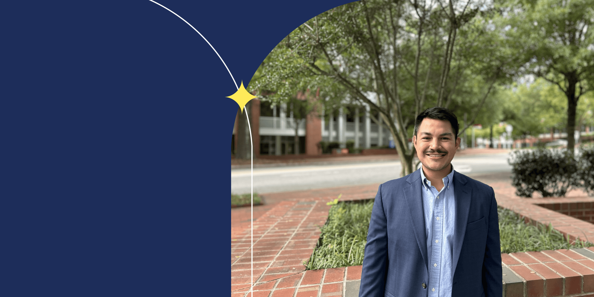 A smiling person facing the camera and standing outside. They are wearing a blue blazer and a blue collared shirt. Behind them, there is a tree.
