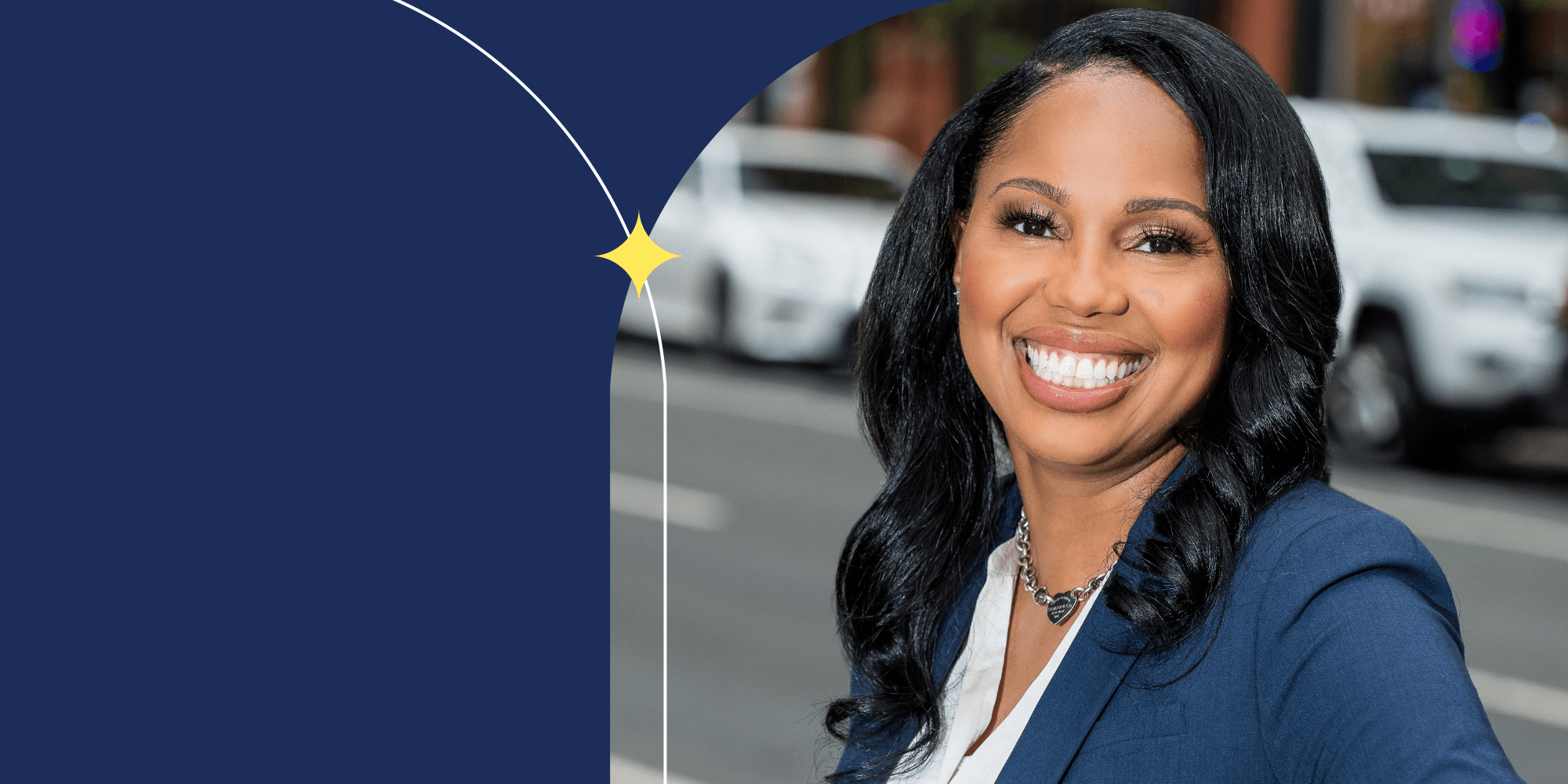 A person facing the camera and smiling. They have long black hair and are wearing a blue blazer. Cars can be seen in background.