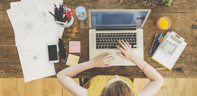 person sitting down at a table working on a laptop with pens, papers, a notebook, and a calculator nearby