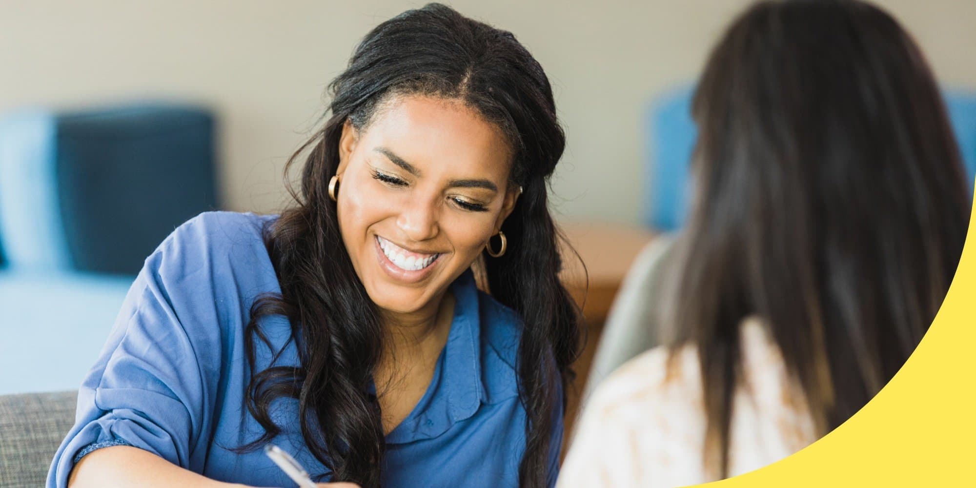 Two people talking in a conference room, one facing the camera smiling and writing with a pen