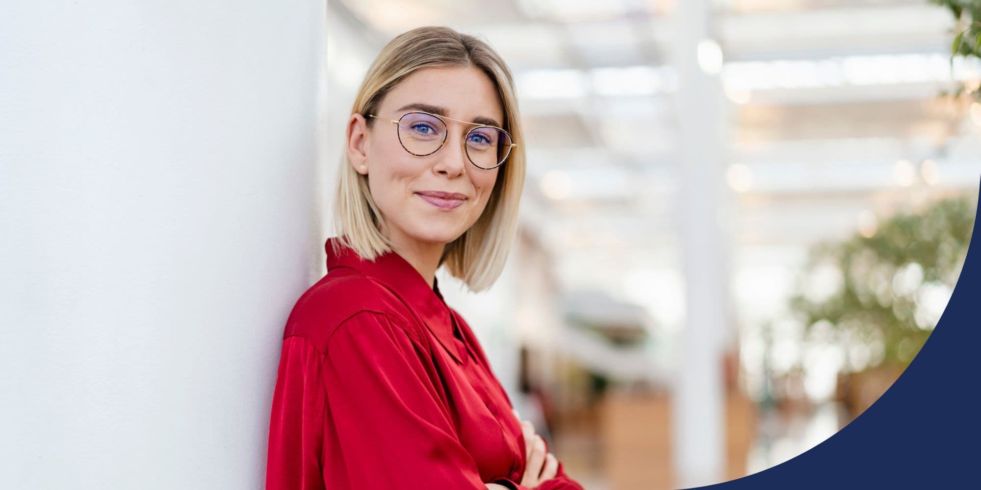 person standing in an office, leaning against a white wall and smiling
