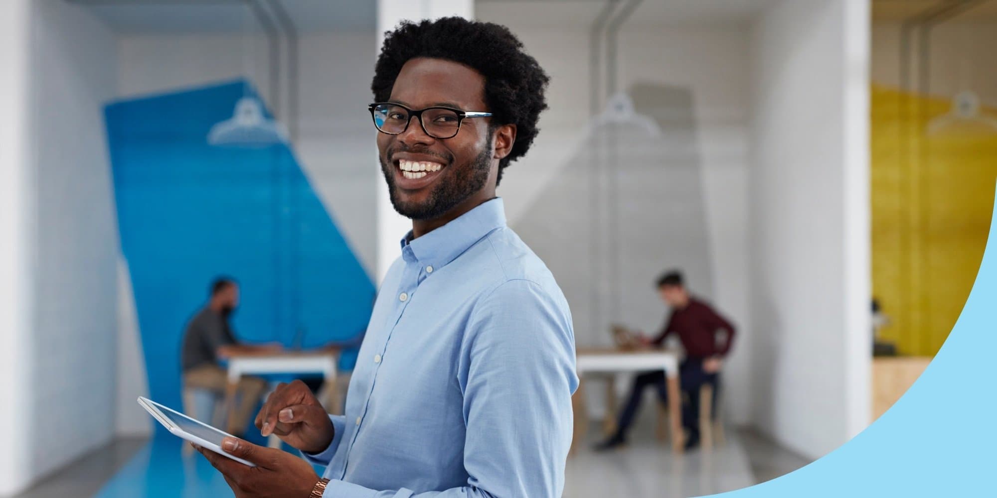 person standing in an office holding a tablet and smiling, with two other colleagues visible in offices behind them